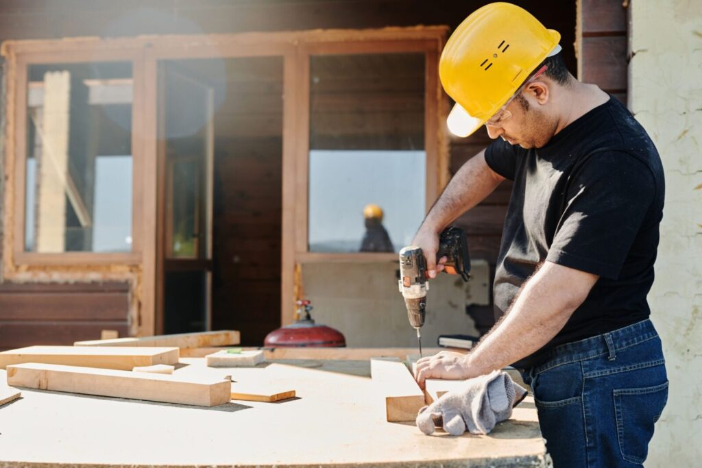 A construction worker with a hard hat uses a power drill on a wooden plank at a renovation site.